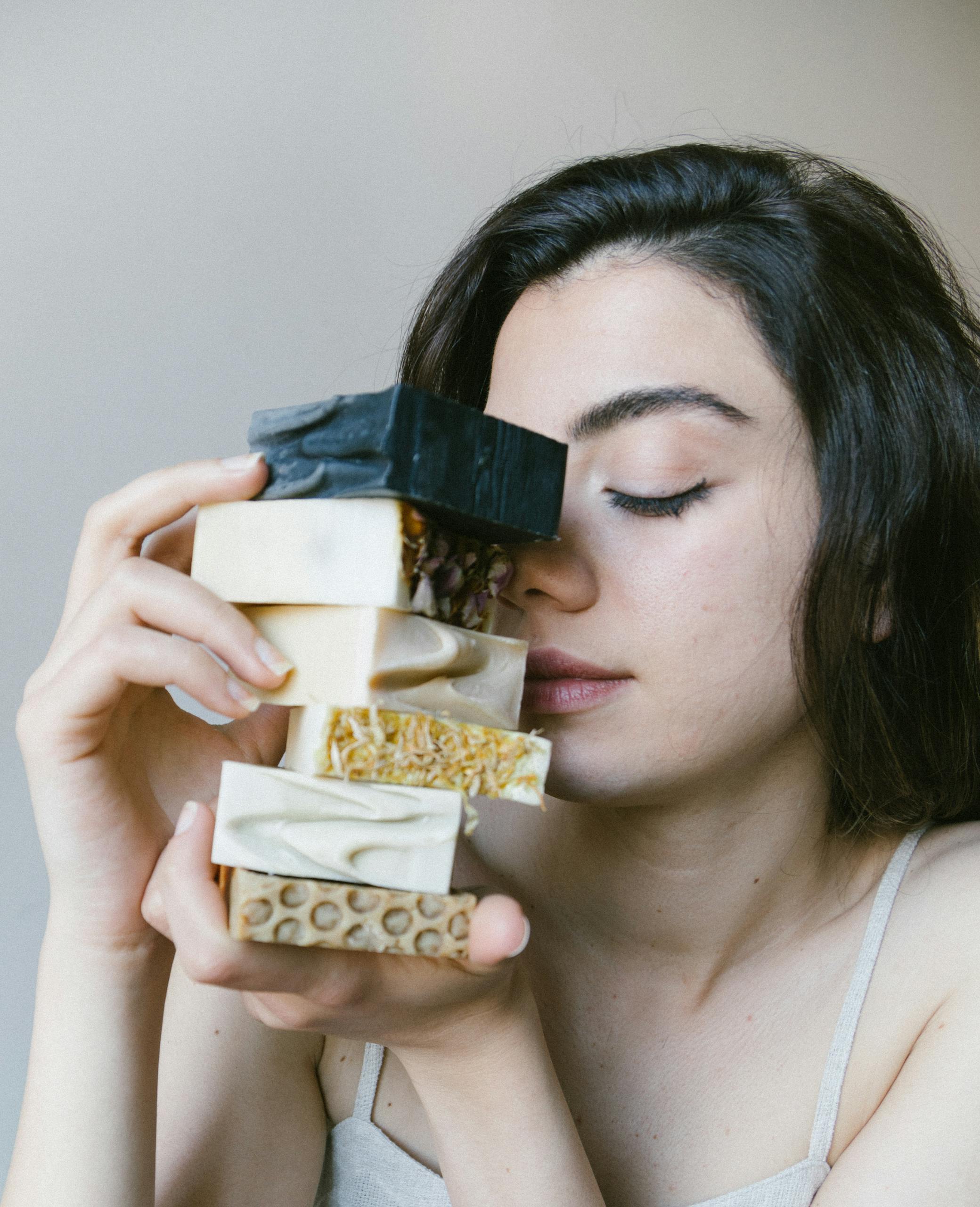 Young woman holding natural soaps with eyes closed, in an indoor setting.